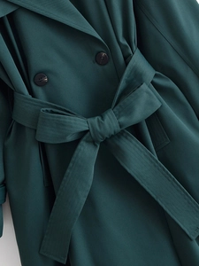 Close-up of dark green trench coat showing twill fabric texture, belt, and black buttons