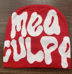 Close-up of red knit beanie featuring white MEA CULPA letters on a dark wood background