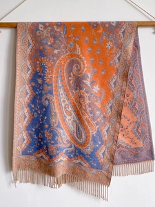 Detailed view of orange and blue paisley cashmere-like scarf with fringe hanging on wood