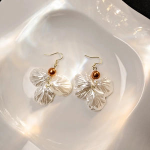 Artistic close-up of shell petal pearl earrings with golden hooks on textured ceramic tray