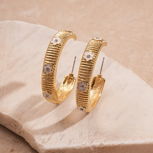Close-up of golden C-shaped hoop earrings with striped texture and white enamel flowers