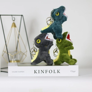 Three colorful plush dinosaur keychains in green, grey and teal stacked on a book