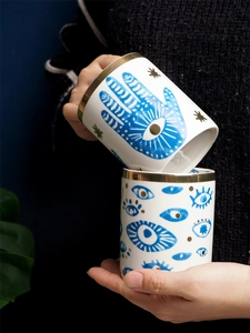 Detail of ceramic mugs featuring blue Hamsa hand and evil eye patterns with gold rim
