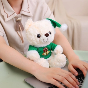 Close-up of a white teddy bear in a green Christmas outfit sitting on a desk with laptop