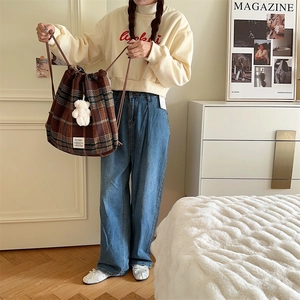 Brown plaid drawstring backpack with white plush charm shown in a cozy bedroom setting