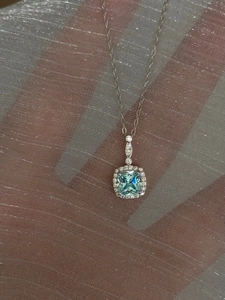 Close-up of a silver necklace with a square light blue gemstone pendant and halo setting