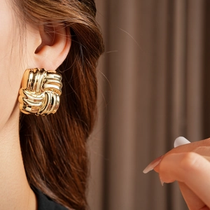 Detail of a square gold metal earring with a woven knot texture on a model's ear