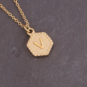 Detail of a gold hexagonal pendant featuring an embossed letter V on a cross chain