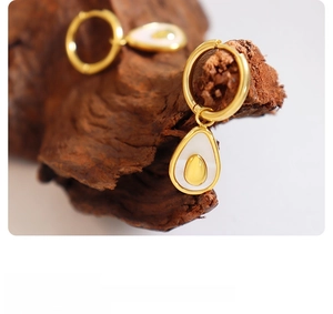 Macro detail of gold avocado-shaped drop earrings with white enamel on a wood surface