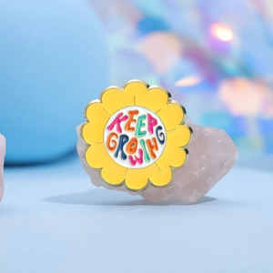 Yellow flower enamel pin with 'KEEP GROWING' text on pink crystal over a pastel background