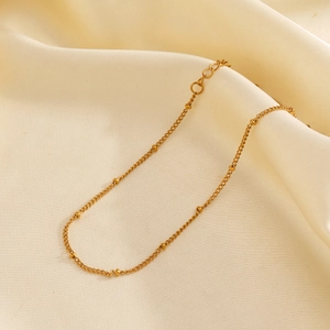 Bracelet de cheville minimaliste en acier doré avec perles posé sur un tissu en soie beige