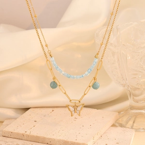 Detail of layered gold butterfly necklace with light blue beads on a beige stone surface