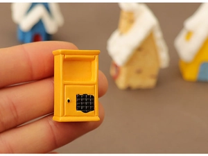Close-up of a miniature yellow mailbox figurine held by fingers for Christmas village scenes
