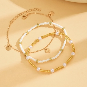 Three-piece bohemian anklet set with gold metal discs and white beaded layering on beige fabric