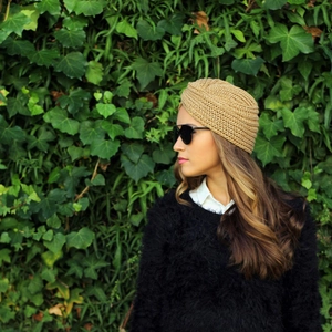 Close-up of a camel knit turban hat with cross-knot detail on a green ivy wall background