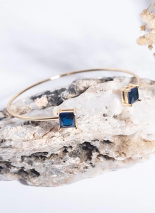 Detail view of gold open bangle with square blue zircons on white natural stone