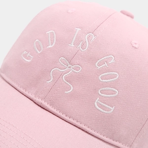 Close-up of white 'GOD IS GOOD' embroidery and bow detail on a pink cotton baseball cap