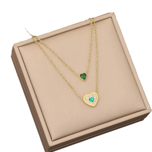 Double-layered gold necklace featuring emerald green heart pendants on beige leather box