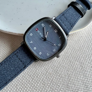 Detail view of square metal case with gray dial and blue suede strap on woven texture