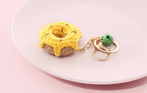 Close-up of a handmade knitted donut keychain in yellow and brown with a green bell