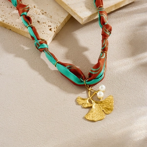 Woven cord necklace with golden ginkgo leaf pendant and white pearl accents on stone background