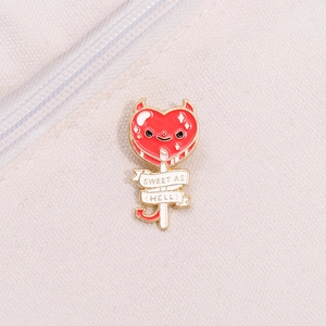 Detailed view of red devil heart lollipop enamel pin on neutral fabric texture