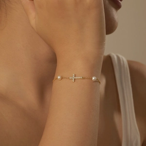Elegant gold cross bracelet with white pearls and zircons worn on a model's wrist