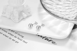 Minimalist silver spherical stud earrings displayed on a white ceramic plate with decor