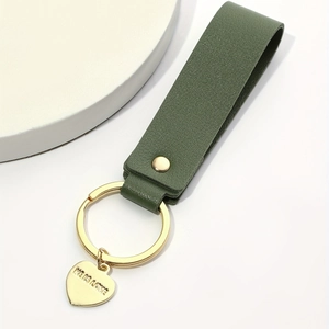 Detail of green leather keychain with gold heart pendant engraved with HE IS LOVE text