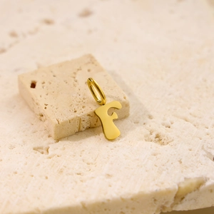 Macro shot of gold stainless steel letter F pendant on a textured beige stone background