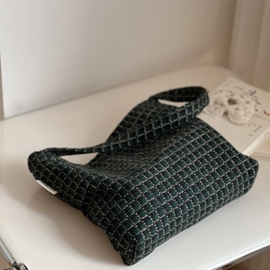Close-up of dark green plaid wool shoulder bag with a woven brand label on white desk