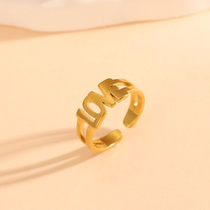 Detail of a golden open ring featuring a metal LOVE letter design on a warm pink background