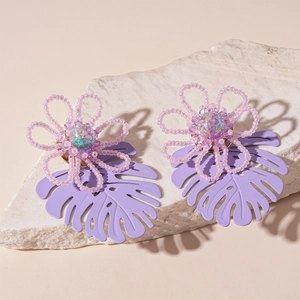 Handmade purple bead flower and monstera leaf drop earrings on a white stone pedestal