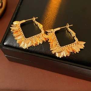 Detailed view of square gold earrings with leaf patterns on a black leather surface