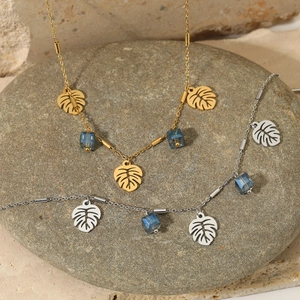 Golden and silver monstera leaf necklace with blue crystal cube on grey stone