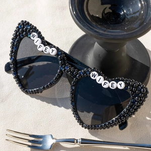 Black heart-shaped bride sunglasses with Wifey beads and pearls on beach background