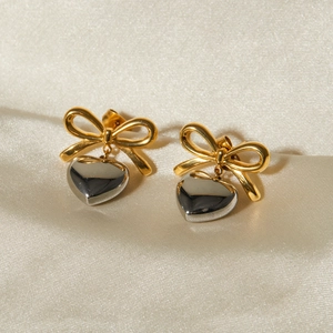 Close-up of gold bow earrings with silver heart pendants on beige satin fabric
