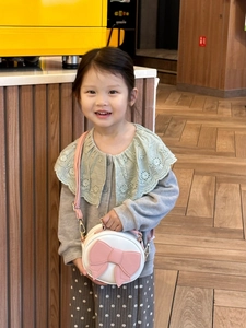 Toddler girl wearing white round crossbody bag with pink bow in a cozy cafe setting