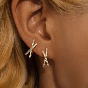 Close-up of golden X-shaped crossover earrings with pavé crystals on a model ear
