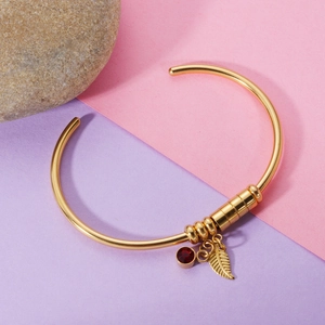 Detail of gold open bracelet with leaf charm and red birthstone pendant on purple background