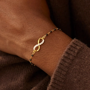 Detail of infinity charm bracelet with white stones and black beads on gold chain