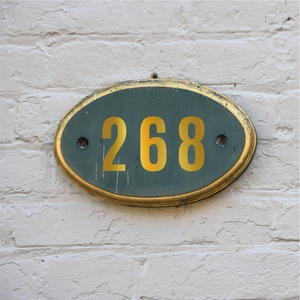 Detail of dark green oval room number plate with gold letters 268 on white brick wall