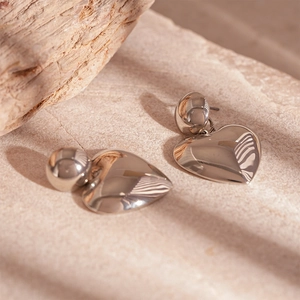 Macro detail of polished silver heart-shaped metal drop earrings on a beige stone surface