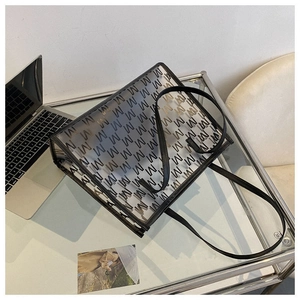 Detail of clear PVC tote bag with M-letter pattern and black leather trim on glass table