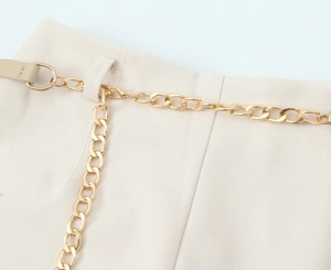 Dettaglio di una cintura a catena in metallo dorato con gancio su pantaloni beige a vita alta