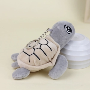 Close-up of a grey plush turtle keychain with a beige shell and metal ring on cream background