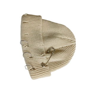 Detailed close-up of beige knitted beanie featuring ripped edges and silver metal safety pin