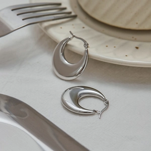 Detail of silver C-shaped steel hoop earrings on linen cloth with a dining scene backdrop