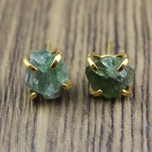 Close-up of raw green crystal stud earrings with gold four-claw setting on wood surface
