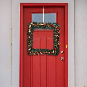 Square green Christmas wreath with red berries hanging on a vibrant red wooden door